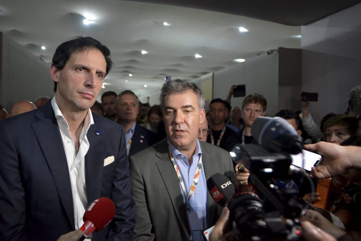 To menn i dress står blant en mengde journalister og mikrofoner og svarer på spørsmål under et innendørs arrangement.