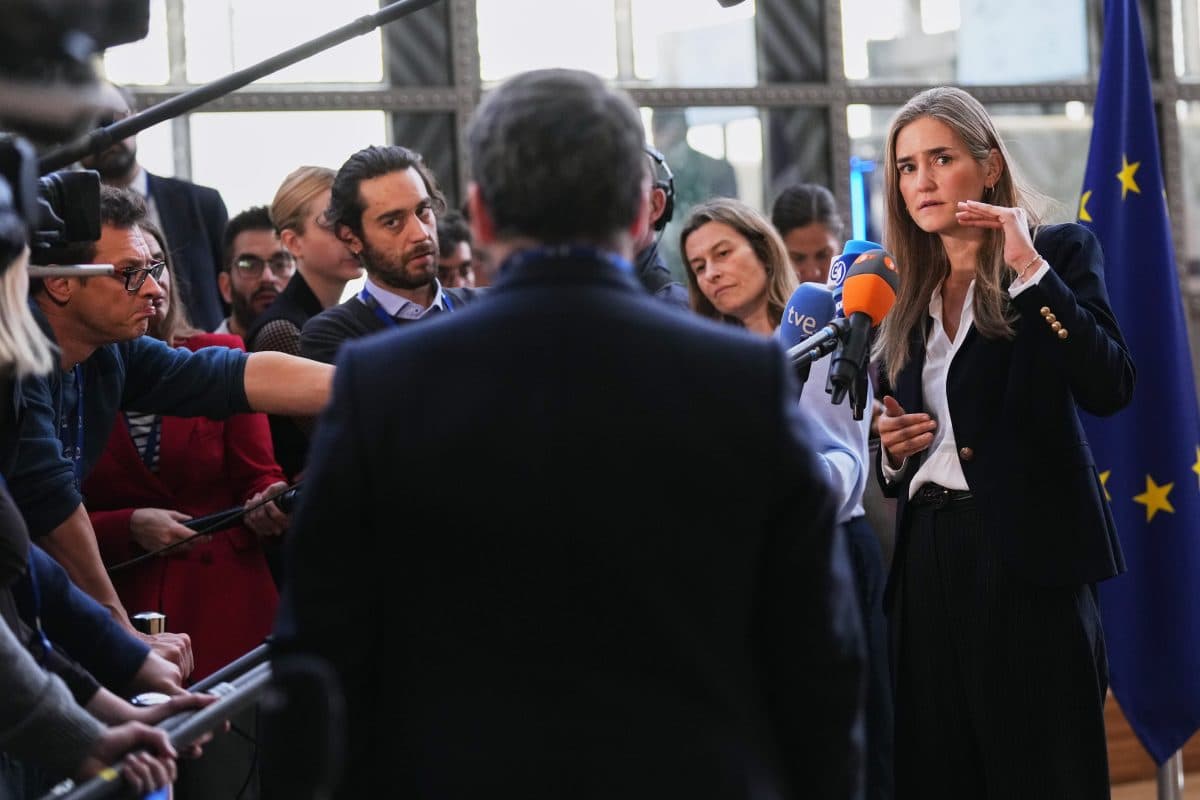 En kvinne i dress taler på en pressekonferanse og gestikulerer med hånden, mens journalister med mikrofoner og kameraer flokker seg rundt henne. Et EU-flagg er synlig i bakgrunnen.