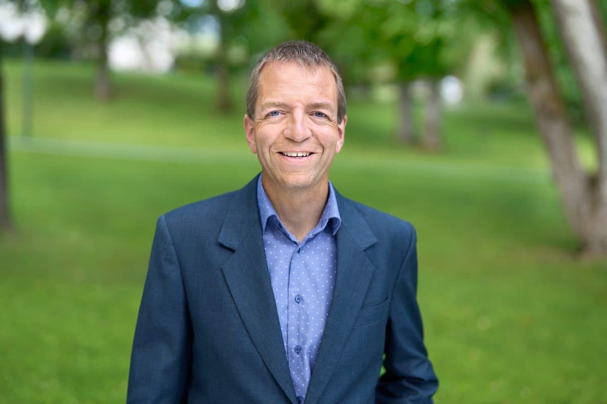 En mann i blå dressjakke og prikkete skjorte står utendørs i en gressbevokst park med trær i bakgrunnen og smiler til kameraet.