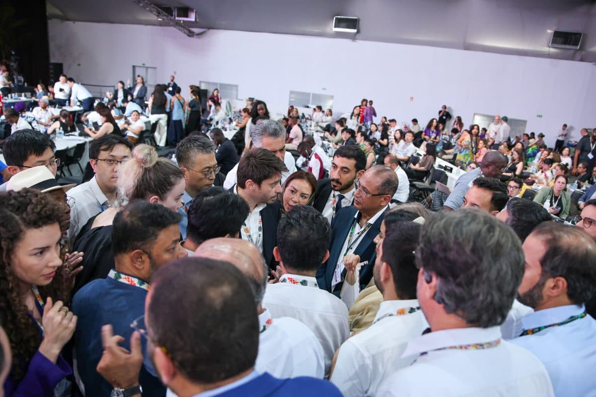 En stor gruppe mennesker, mange med nøkkelbånd, står tett sammen og diskuterer i en travel konferansesal.