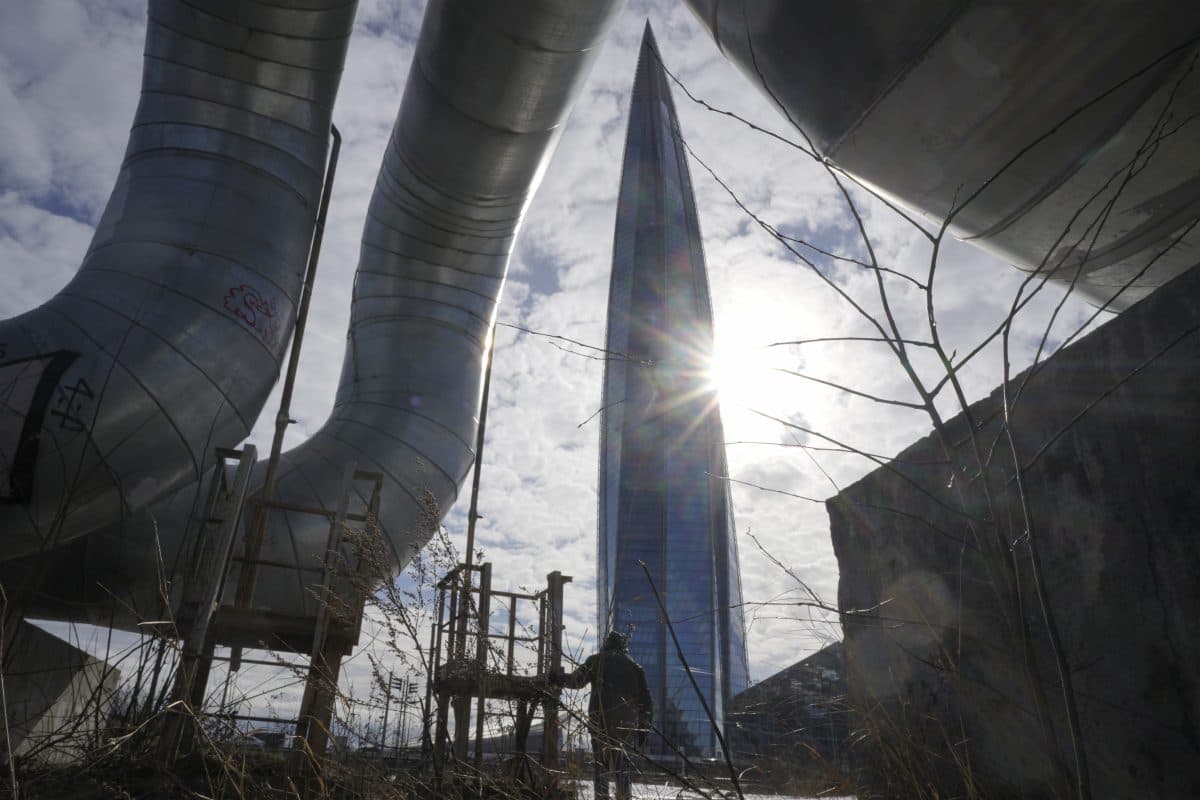 En person står i nærheten av store industrirør med en høy, spiss skyskraper og solen synlig i bakgrunnen.