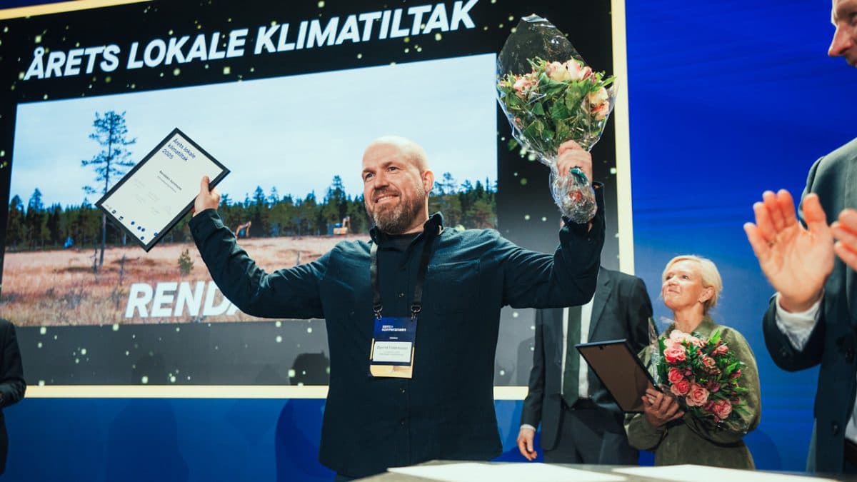 En mann med diplom og bukett står på scenen foran en storskjerm med teksten "ÅRETS LOKALE KLIMATILTAK", mens folk rundt ham applauderer.