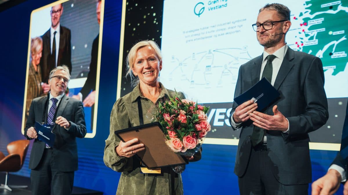 Tre personer i formelle antrekk står på scenen; kvinnen foran holder en bukett og en plakett, mens de to mennene holder mapper. I bakgrunnen skimtes et presentasjonsbilde.