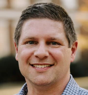 En mann med kort, brunt hår og lys hudfarge smiler mot kameraet, iført rutete skjorte. Bakgrunnen er svakt uskarp.