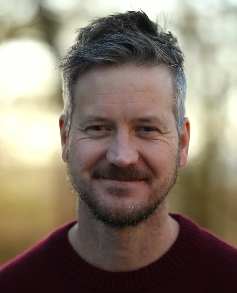 En mann med kort, grått hår og skjegg smiler lett, iført en rødbrun genser, med en uskarp utendørs bakgrunn.