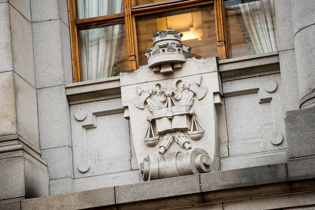 Steinrelieff med et våpenskjold med økser, skjell og skriftruller på ytterveggen av en grå steinbygning, under et vindu med gardiner.