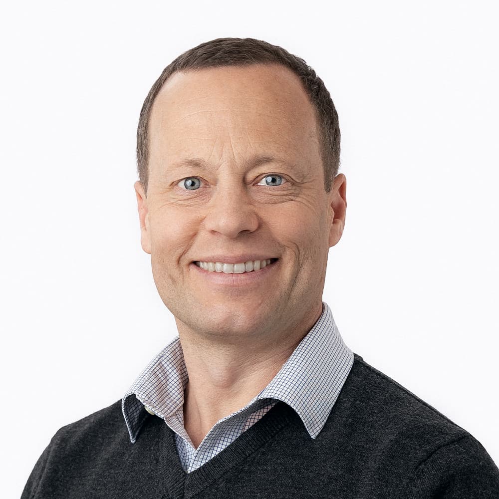 En mann med kort, brunt hår smiler mot kameraet. Han er iført en grå genser over en skjorte med krage mot en ensfarget hvit bakgrunn.