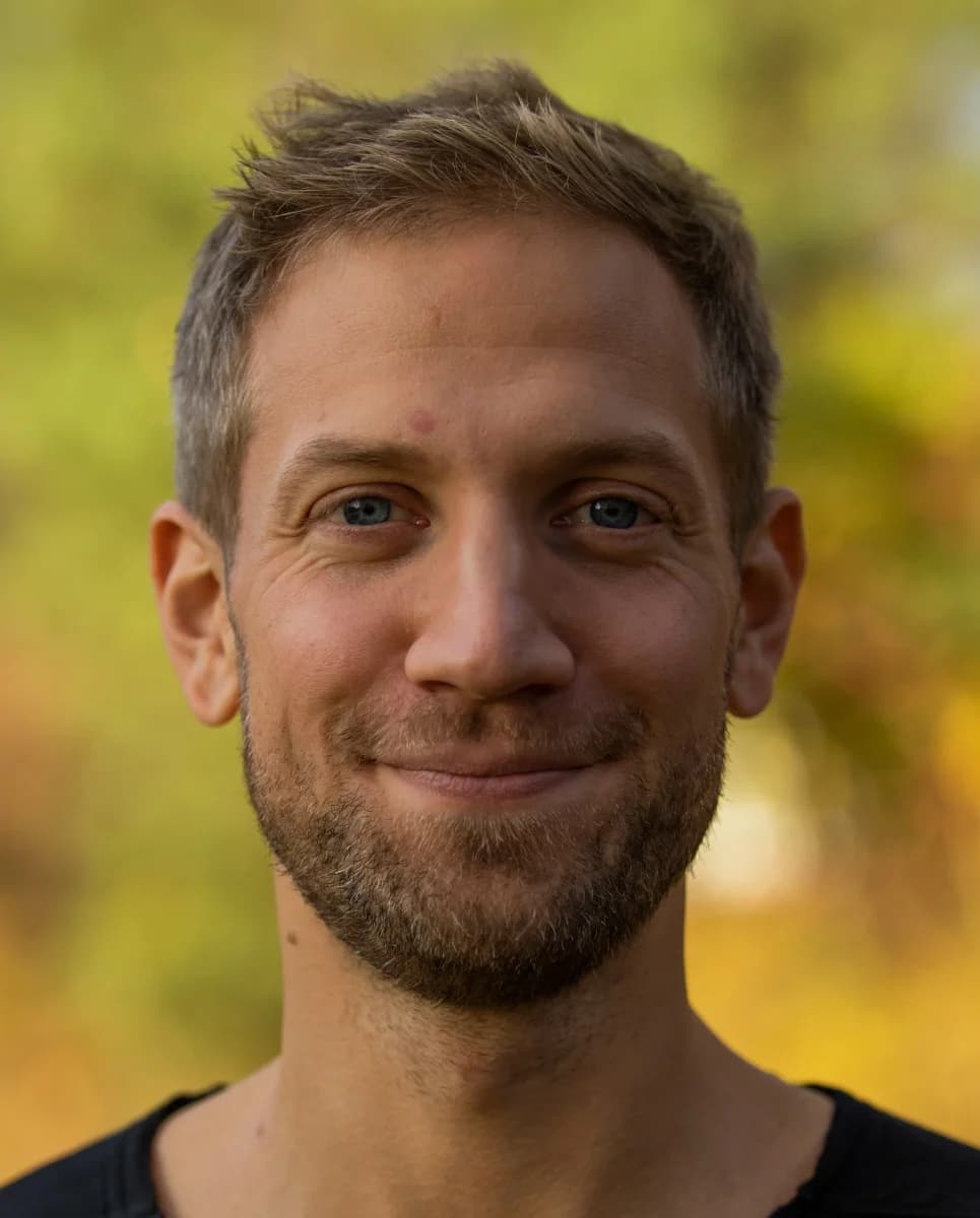 En mann med kort, lysebrunt hår og trimmet skjegg smiler til kameraet utendørs, med en uskarp grønn og gul bakgrunn.