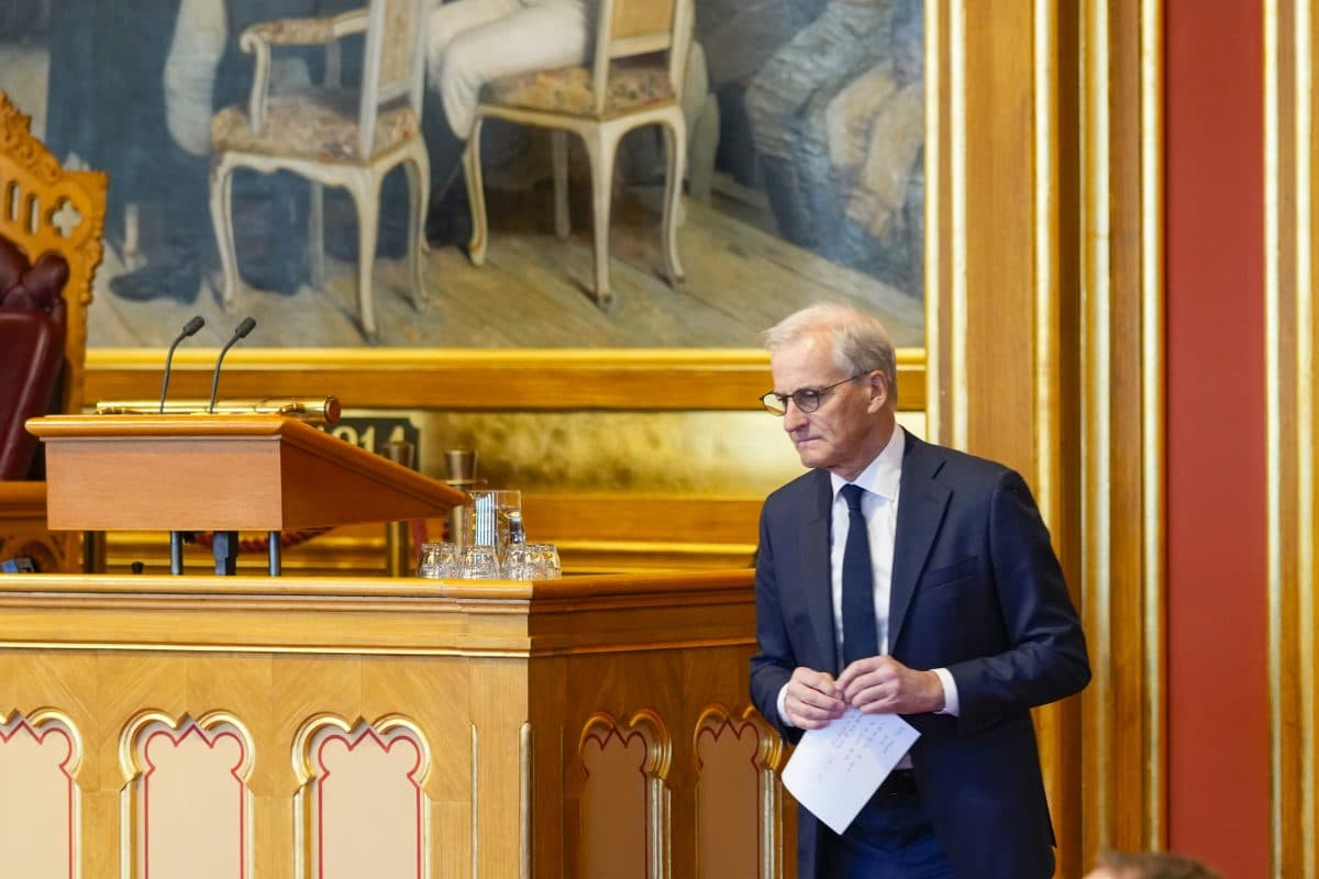 En mann i dress står ved et podium i et formelt rom med utsmykkede tredetaljer og et stort maleri i bakgrunnen.