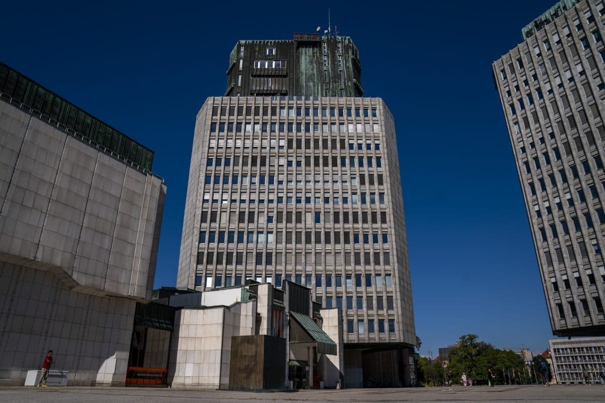 En høy, rektangulær kontorbygning med en fasade av glass og betong står mellom to andre moderne bygninger under en klarblå himmel.