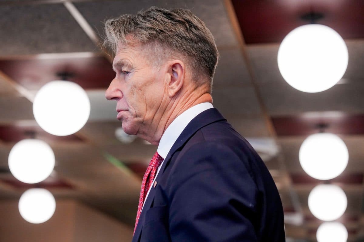 En mann i dress og rødt slips er avbildet i profil, stående innendørs med runde taklamper i bakgrunnen.