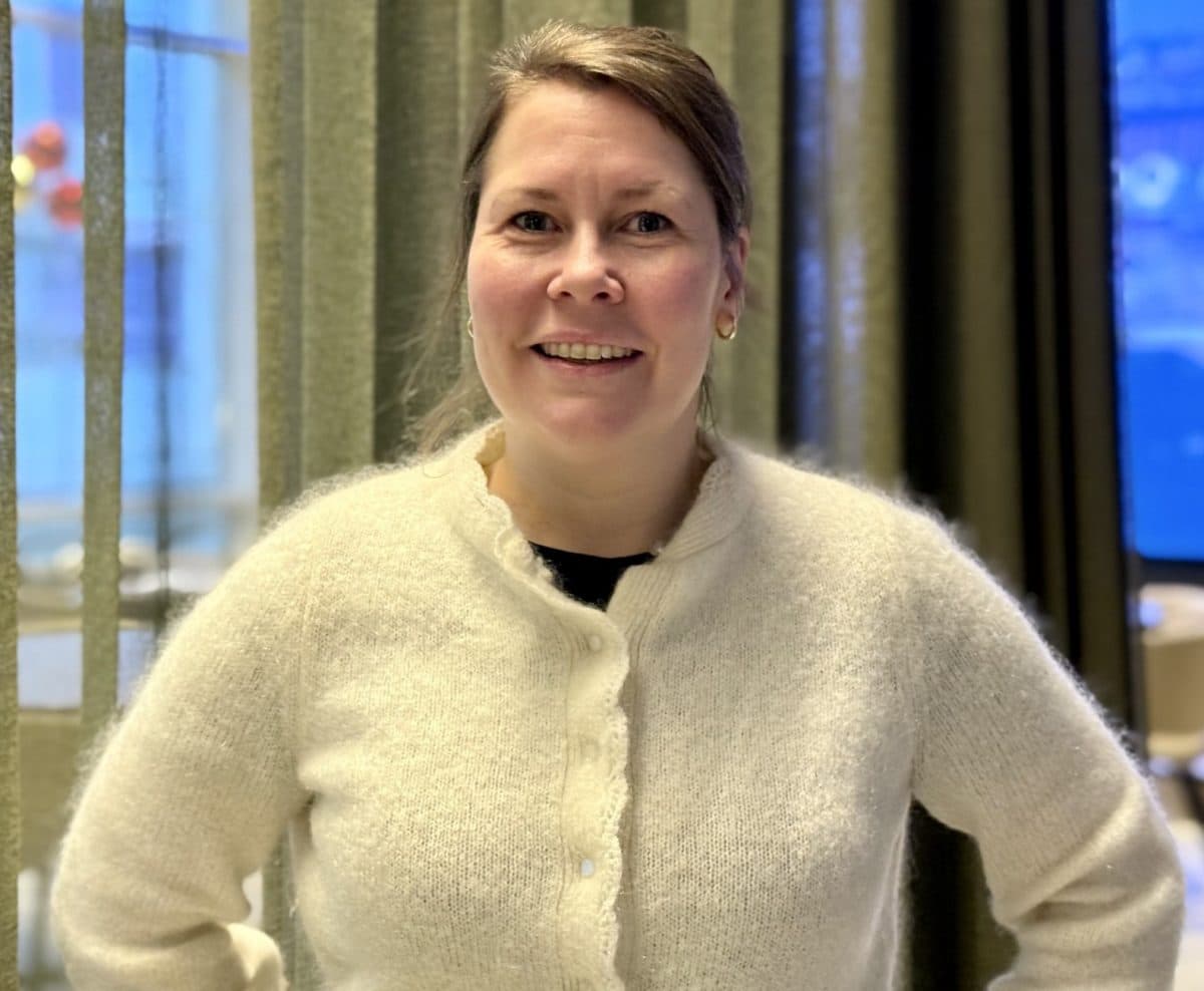 En kvinne med lysebrunt hår, iført en hvit, lodden cardigan, står innendørs foran grønne gardiner og smiler til kameraet.