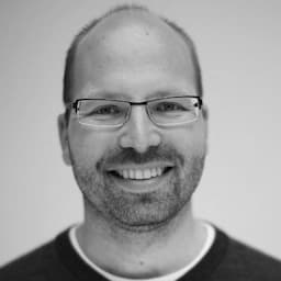 En mann med briller, skjegg og vikende hårfeste smiler til kameraet mot en ensfarget bakgrunn.