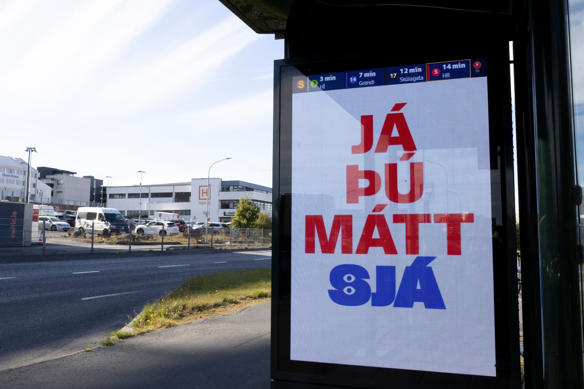 A digital bus stop display shows "JÁ ÞÚ MÁTT" in red text and "SJÁ" in blue text, with a street and buildings in the background.