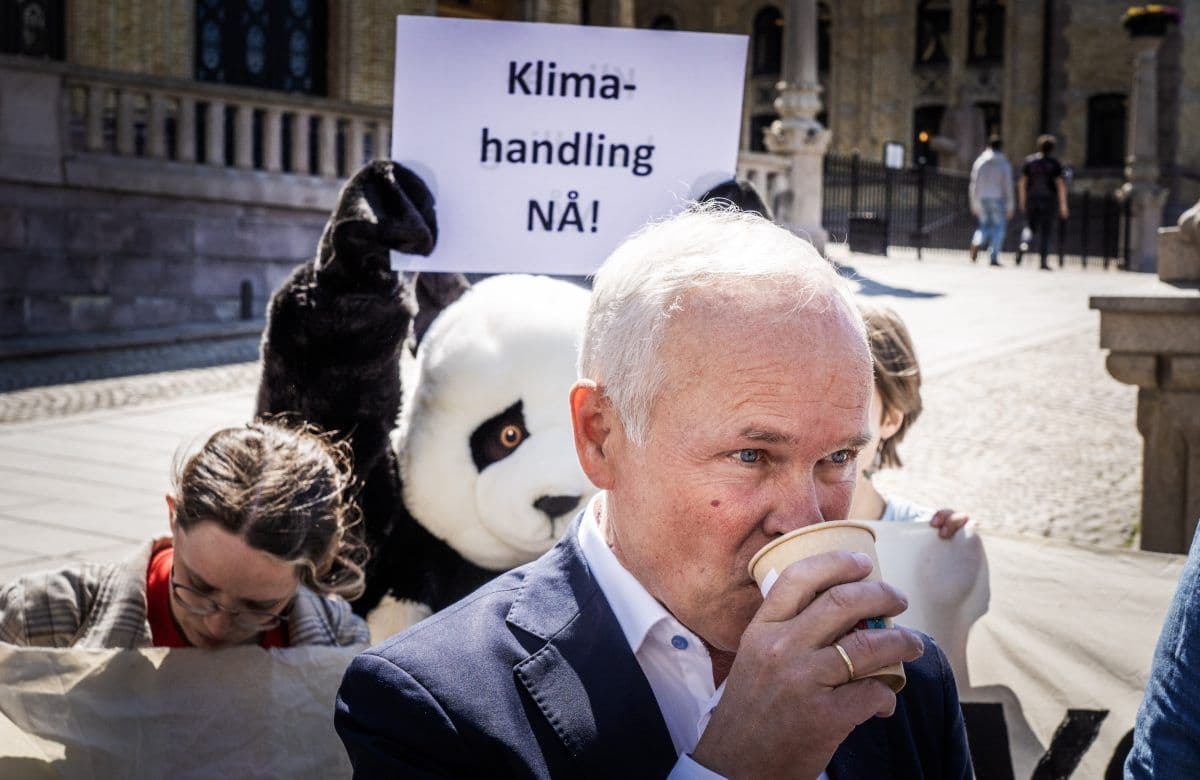 En mann drikker av en pappkopp mens en person i pandadrakt holder et skilt med teksten «Klima-håndtering NÅ!» under en utendørs klimaprotest.
