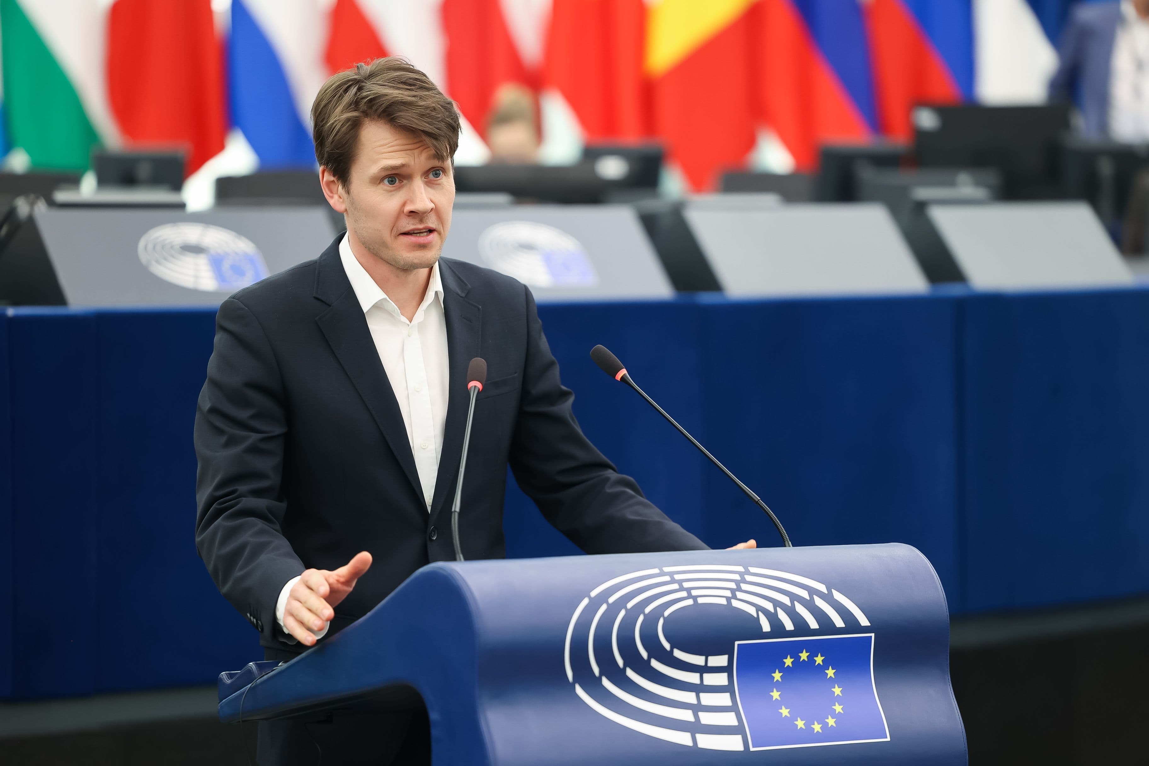 En mann i dress taler fra en talerstol med EU-flagget og Europaparlamentets logo, i en sal med internasjonale flagg.
