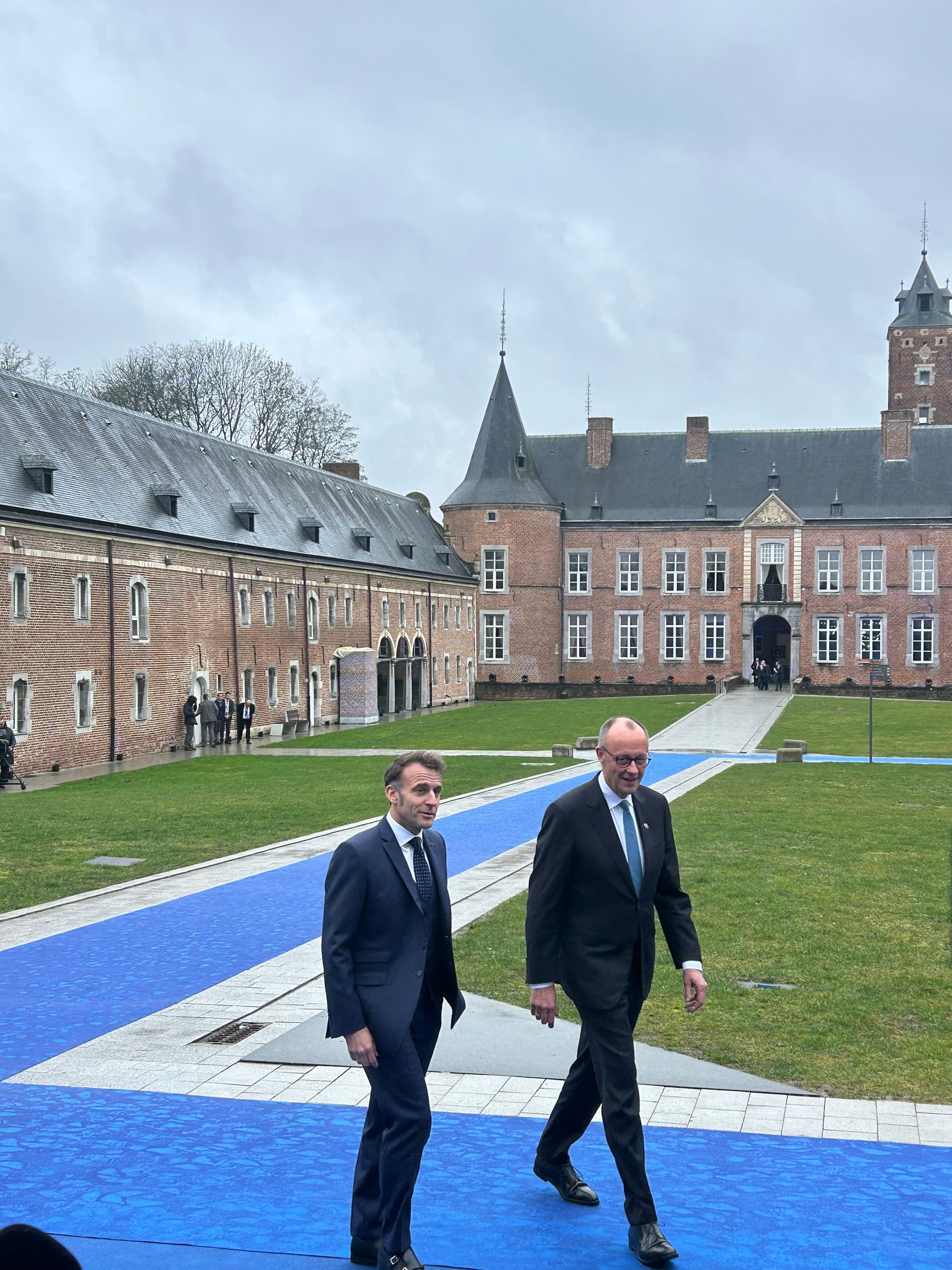 Emmanuel Macron and another man walk on a blue carpet in front of a large brick chateau.