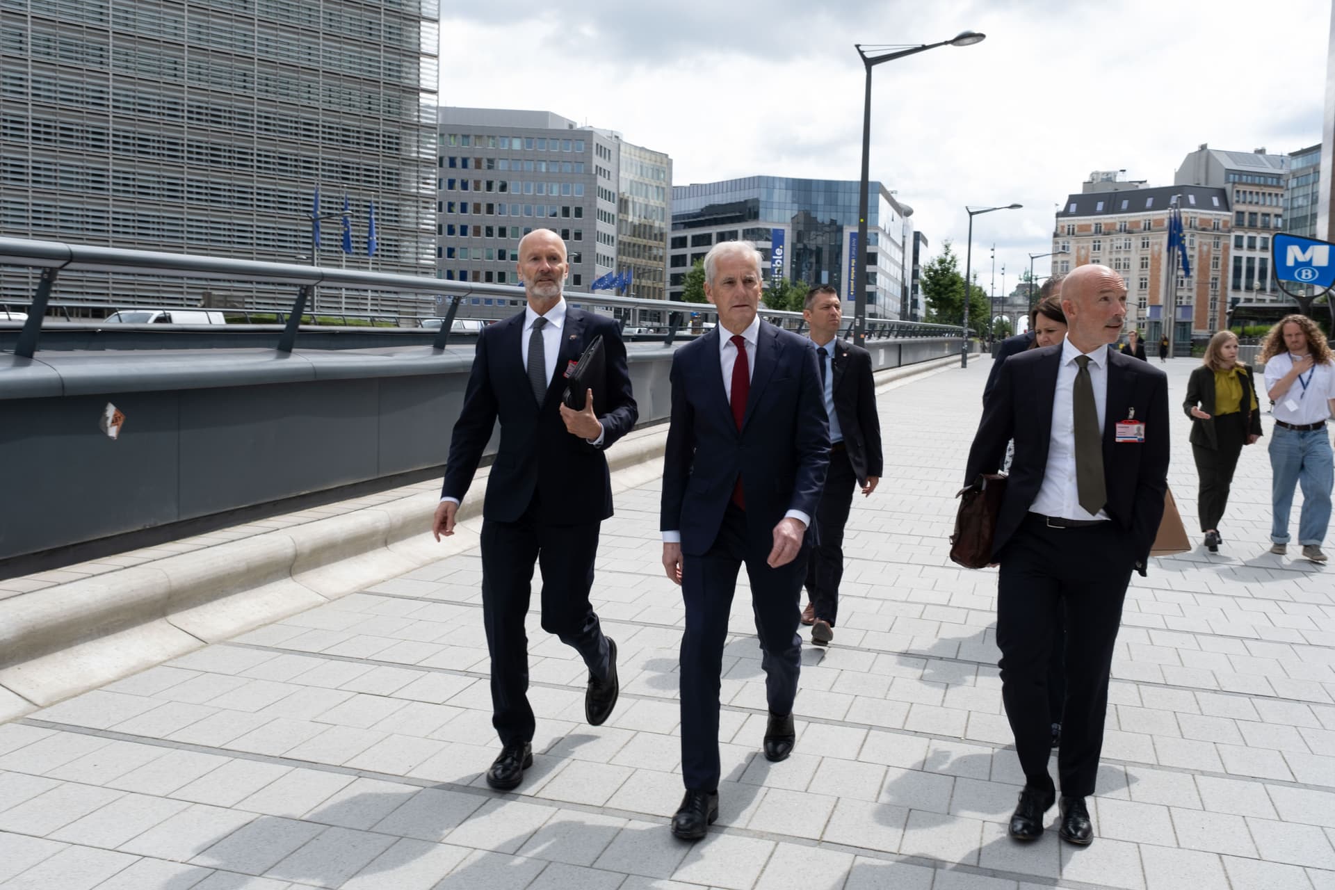 A group of men in dark suits walk on a paved walkway through a modern city district.