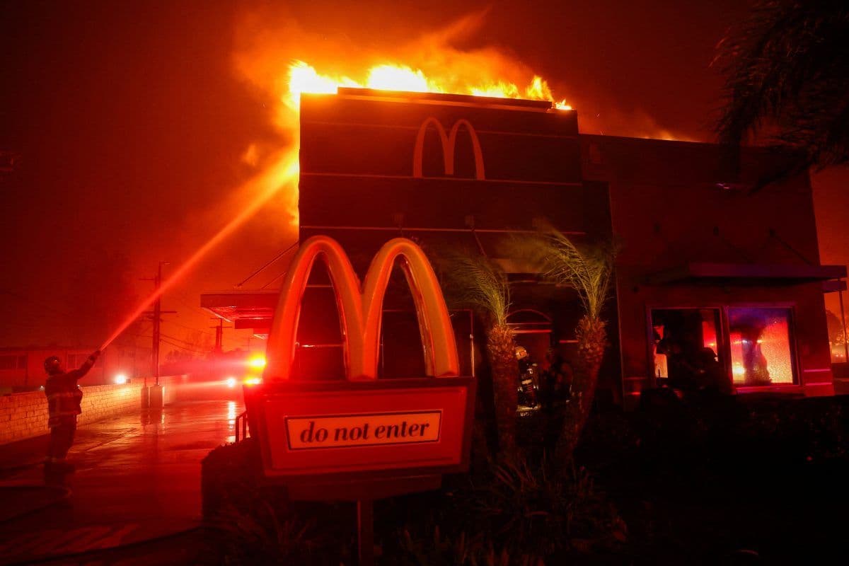 En McDonald's-restaurant står i brann om natten, med flammer fra taket. Brannmannskaper spruter vann fra slanger for å få kontroll over brannen. Et "adgang forbudt"-skilt er synlig foran.
