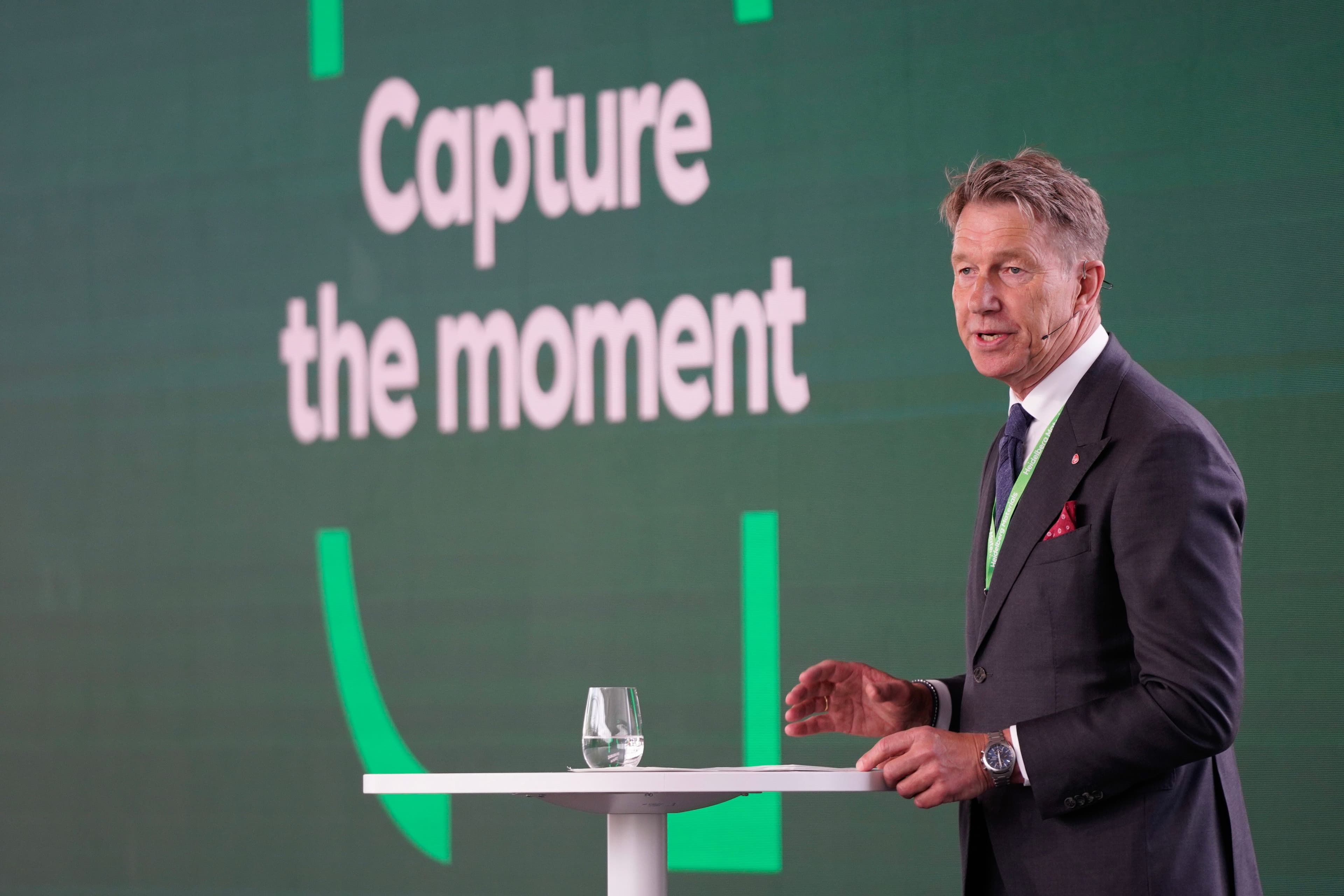 Mann i dress som taler fra en talerstol, med «Capture the moment» vist på en grønn skjerm bak ham.