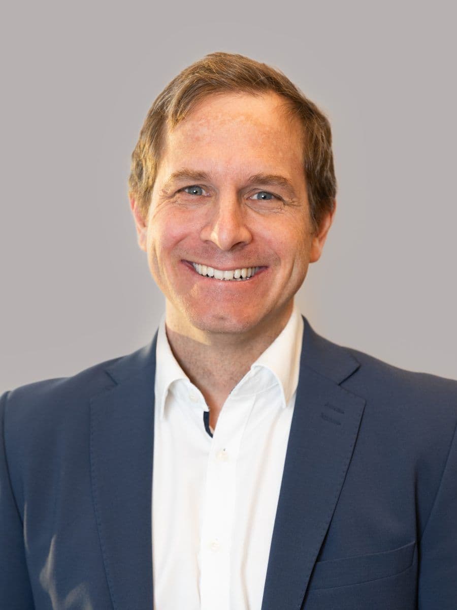 En mann iført blå dressjakke og hvit skjorte smiler til kameraet mot en enkel, lysegrå bakgrunn.