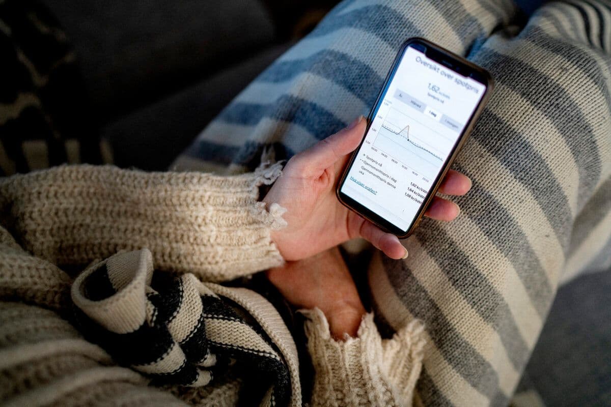 Person som sjekker strømprisen på en smarttelefon mens hun slapper av på en sofa med et stripete teppe.