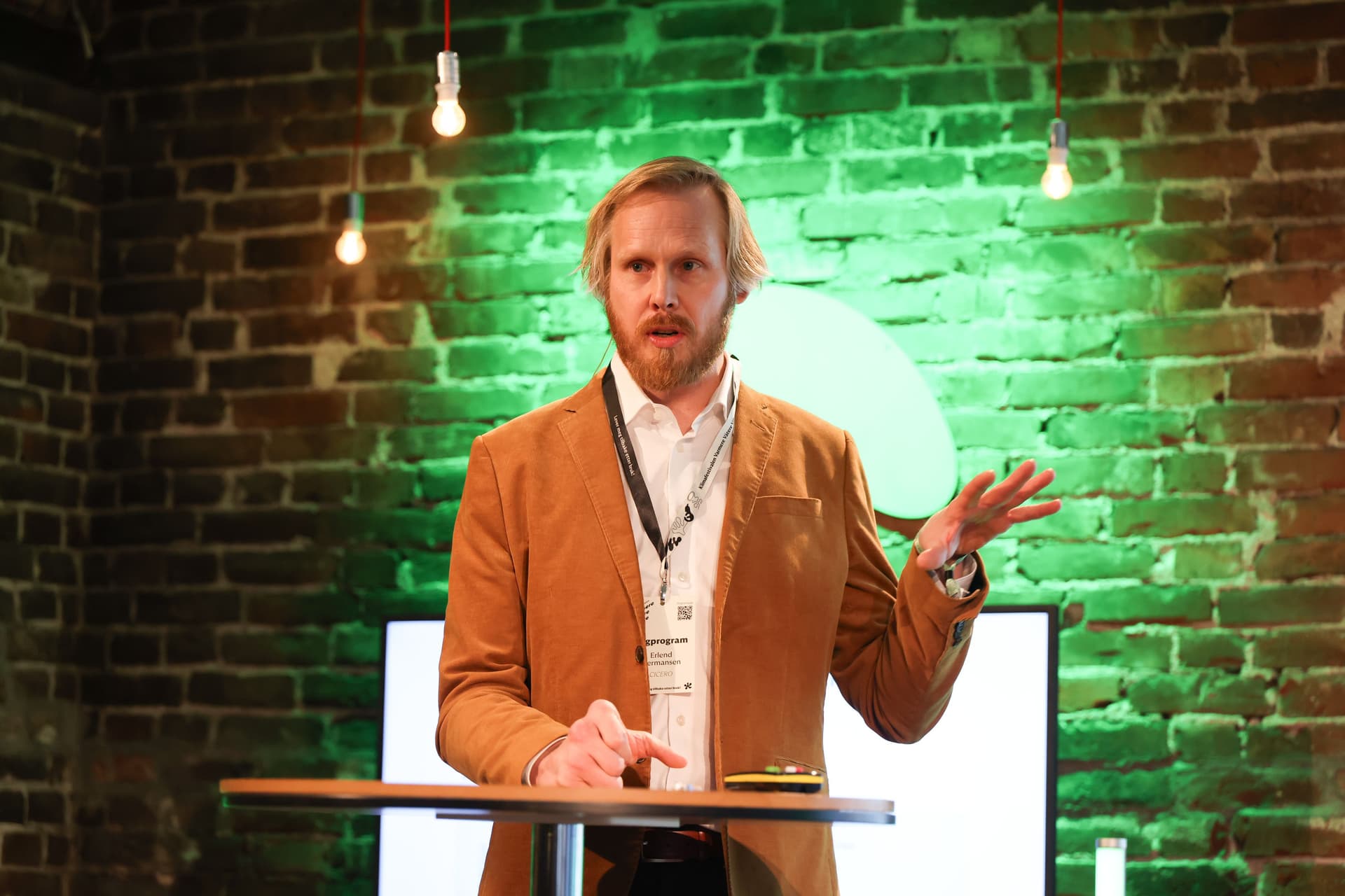 En skjeggete mann i brun jakke gestikulerer mens han presenterer foran en grønt opplyst murvegg.