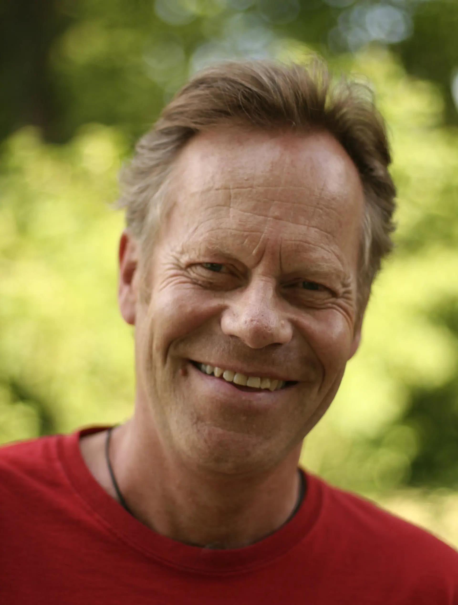 En smilende mann med kort hår, rød t-skjorte står foran grønne blader i bakgrunnen.