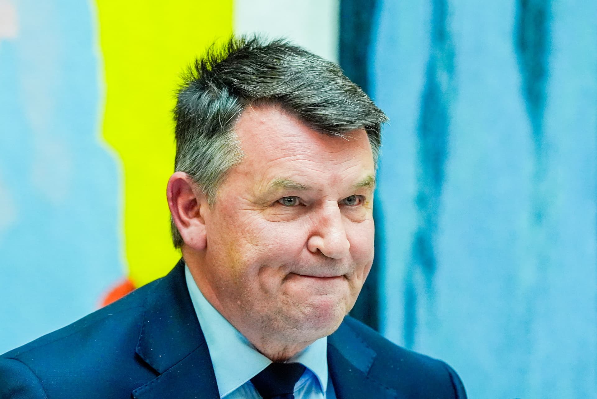 Tor Mikkel Wara i dress ser til høyre med et svakt smil, foran en fargerik, uskarp bakgrunn.