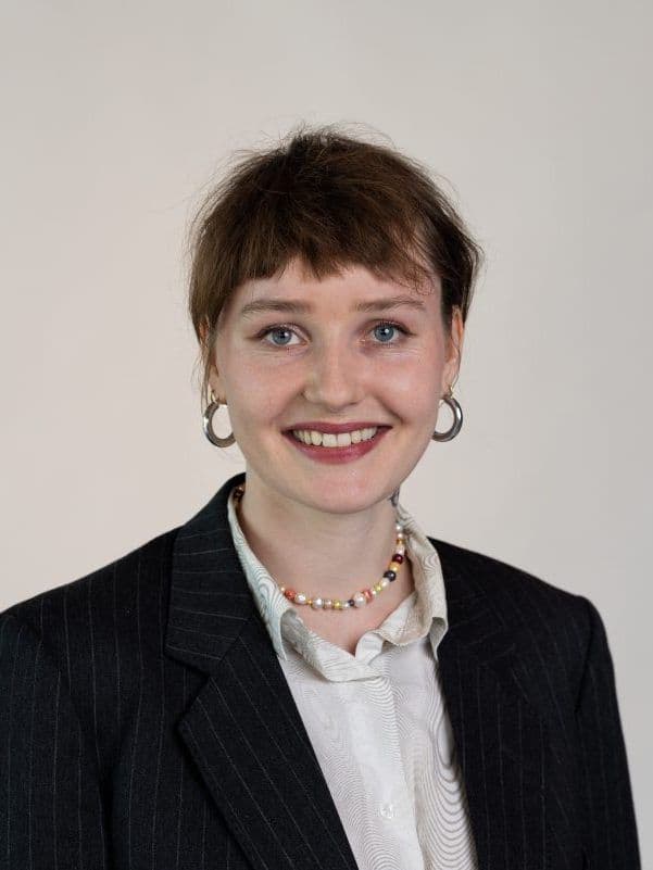 Person med kort hår og øredobber, iført stripet dressjakke og skjorte med krage, smilende mot en vanlig bakgrunn.