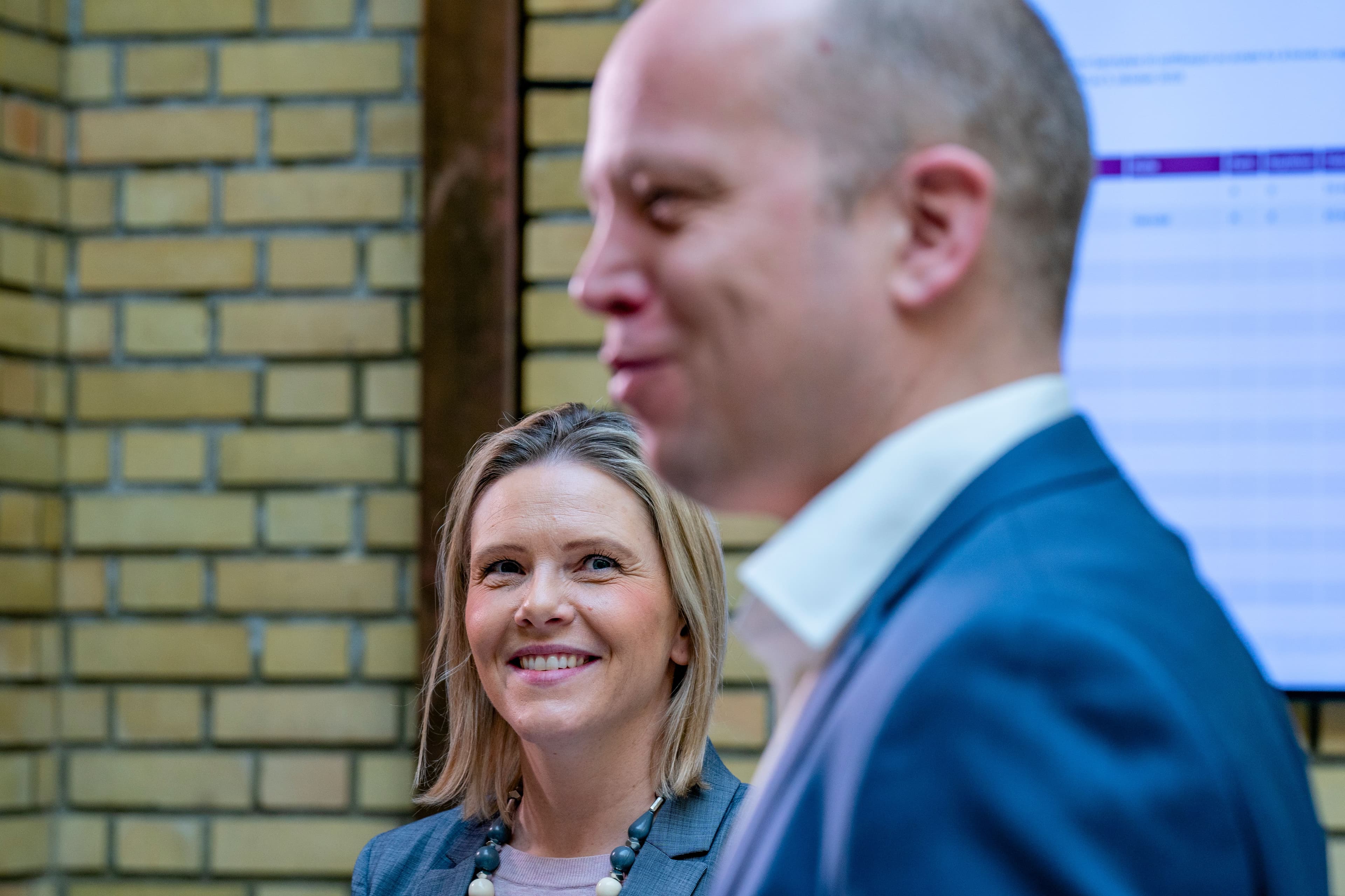 En smilende blond kvinne ser fremover, med en uskarp mannsprofil i forgrunnen.