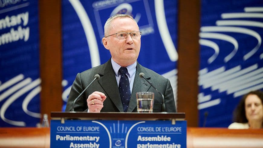 A man in a suit and glasses speaks at a podium with "Council of Europe Parliamentary Assembly" visible.