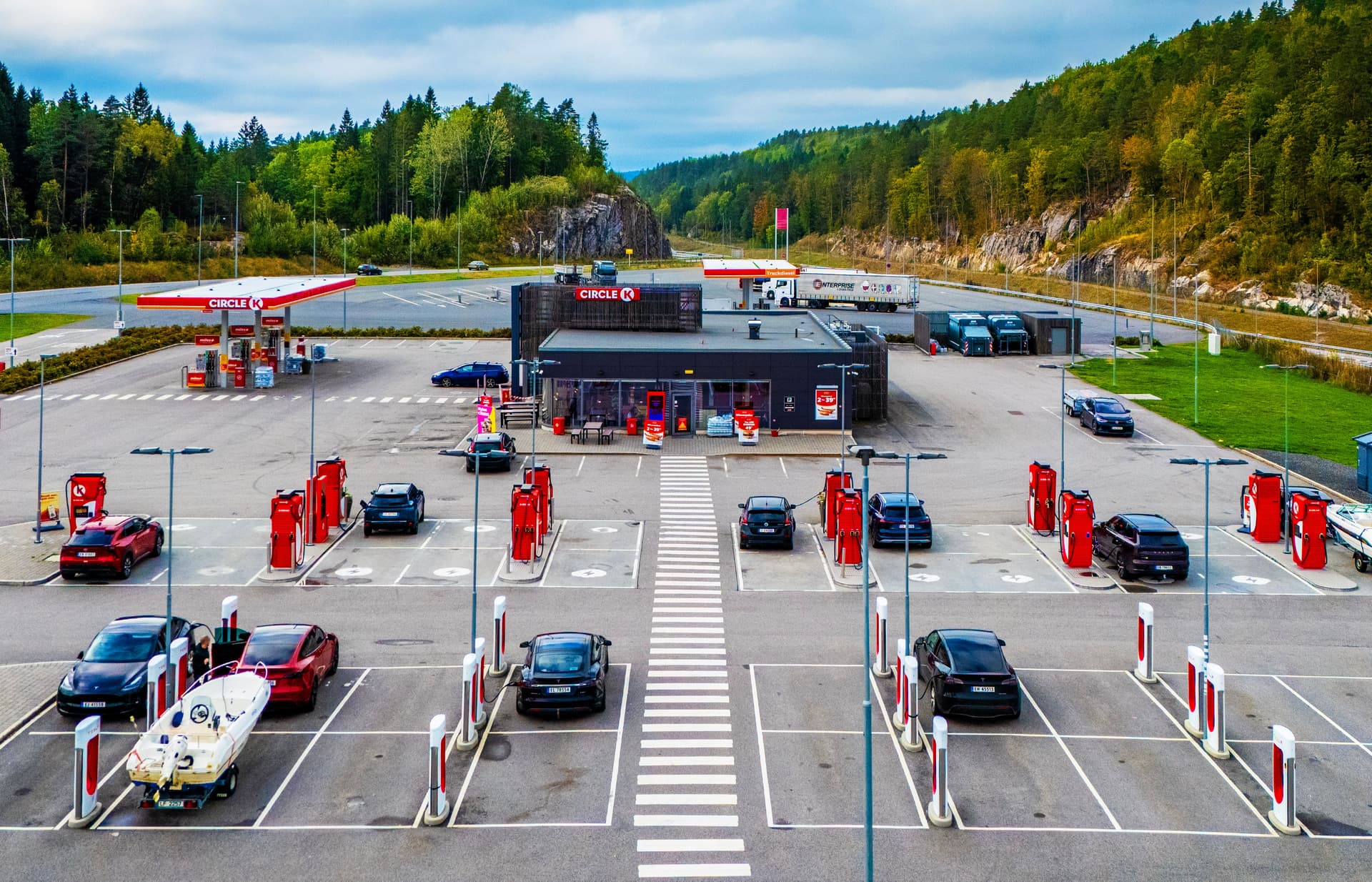 Flyfoto av en rasteplass langs veien med en Circle K-bensinstasjon, en storkiosk og flere røde ladestasjoner for elbiler. Flere biler står parkert, noen til lading, og én har en båthenger med båt, med skog i bakgrunnen.