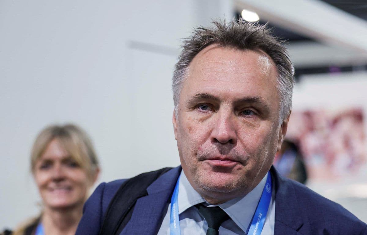 En mann i dress og slips med snor går innendørs. En kvinne er synlig i bakgrunnen og smiler.