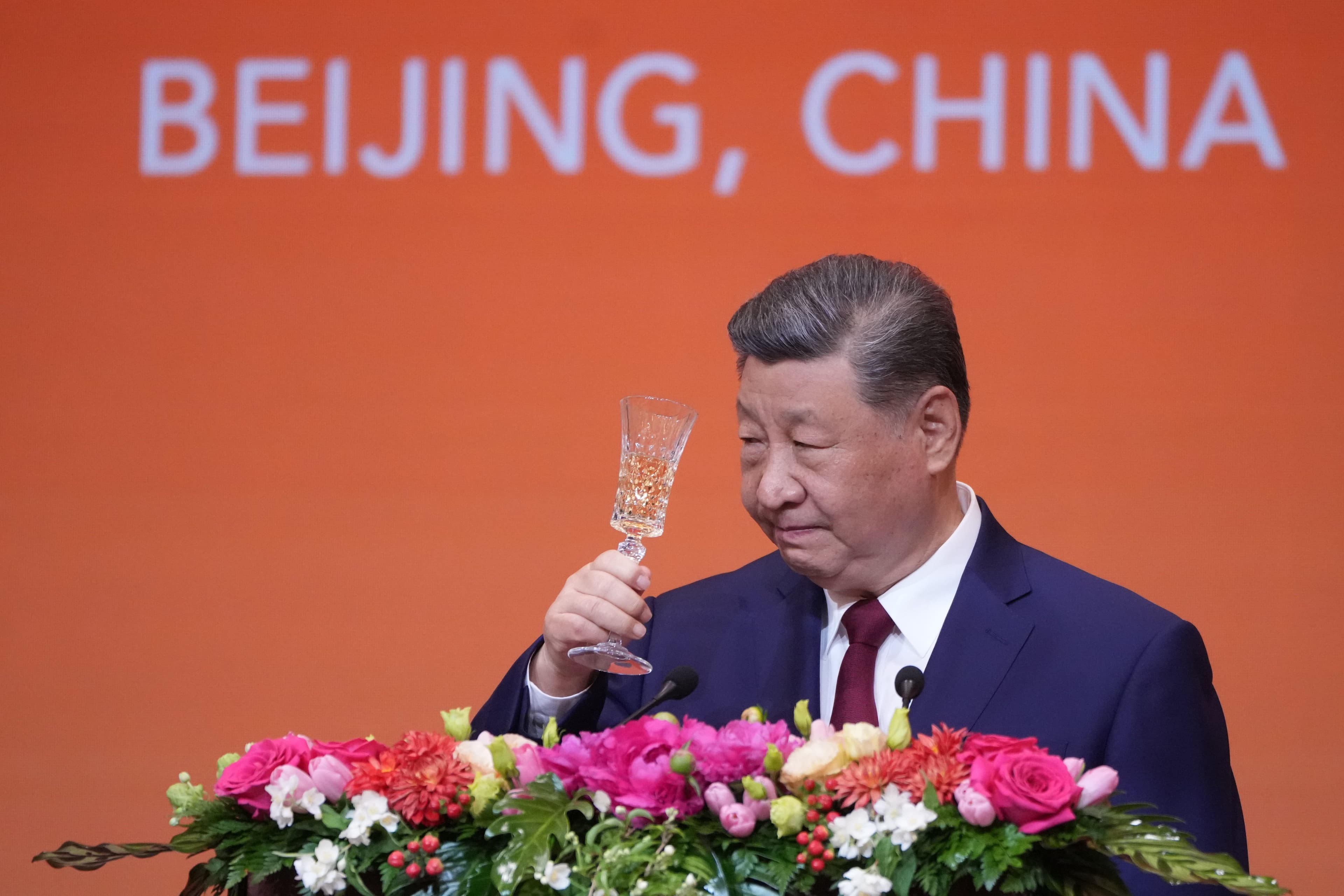 Xi Jinping holder et champagneglass ved en talerstol med blomster, foran et oransje banner med teksten «BEIJING, CHINA».
