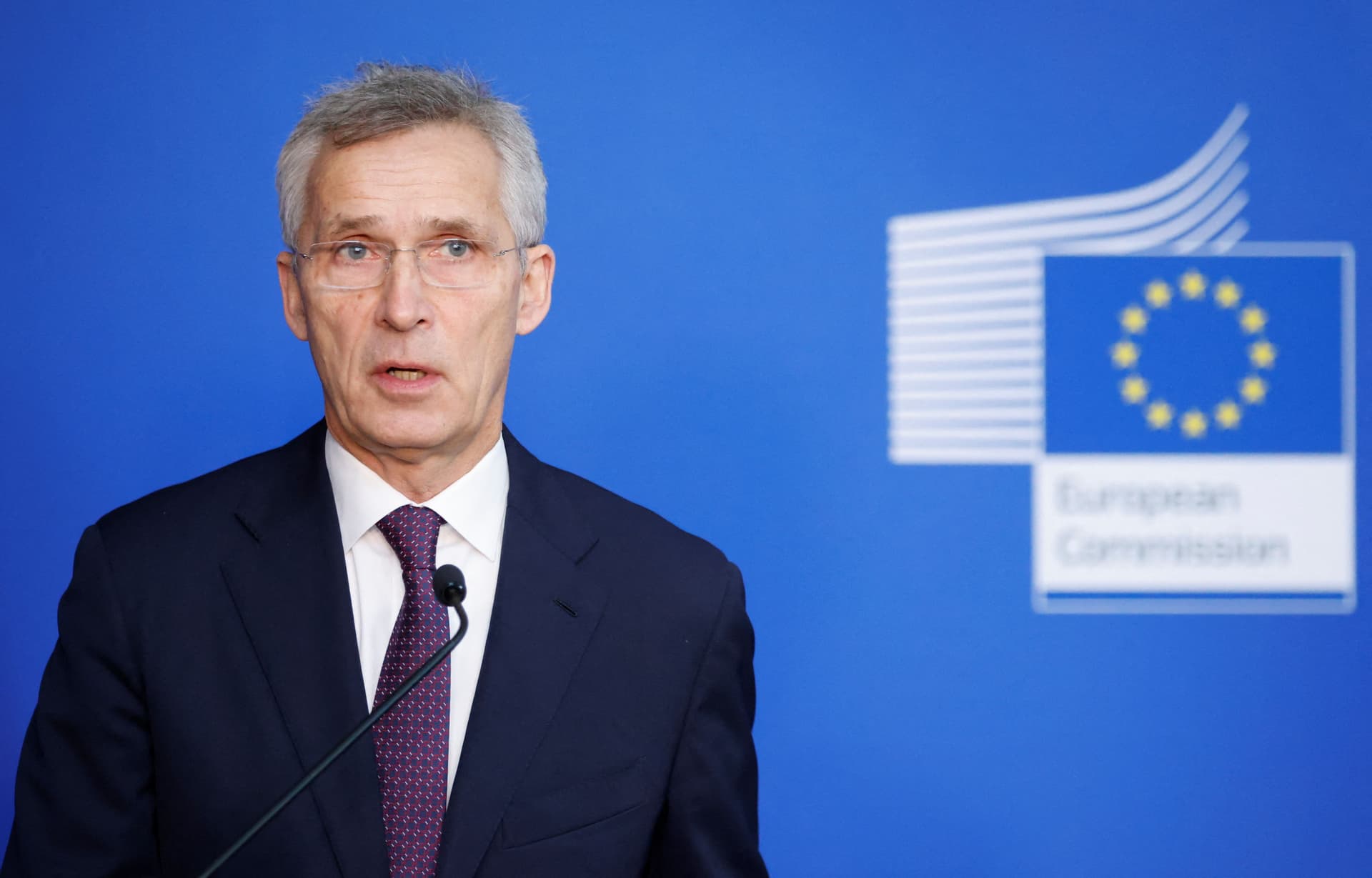 Jens Stoltenberg speaking at a podium with the European Commission logo in the background.