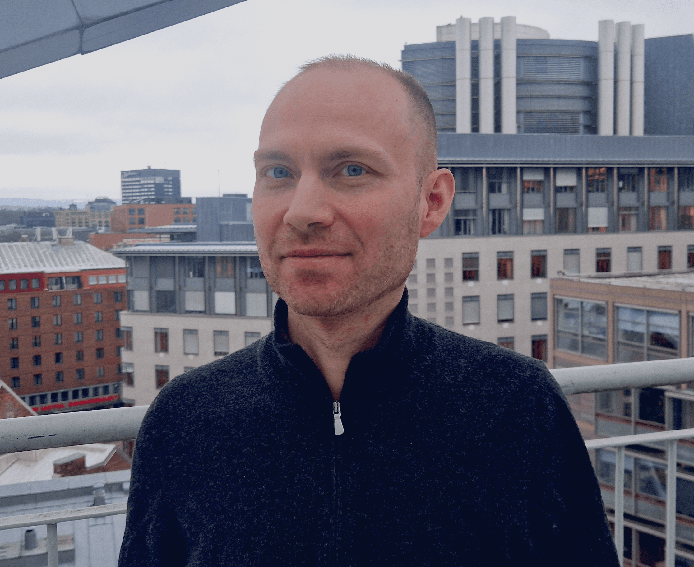 En mann med kort, mørkt hår og blå øyne, iført en mørkegrå genser, smiler svakt på en takterrasse med utsikt over en by.
