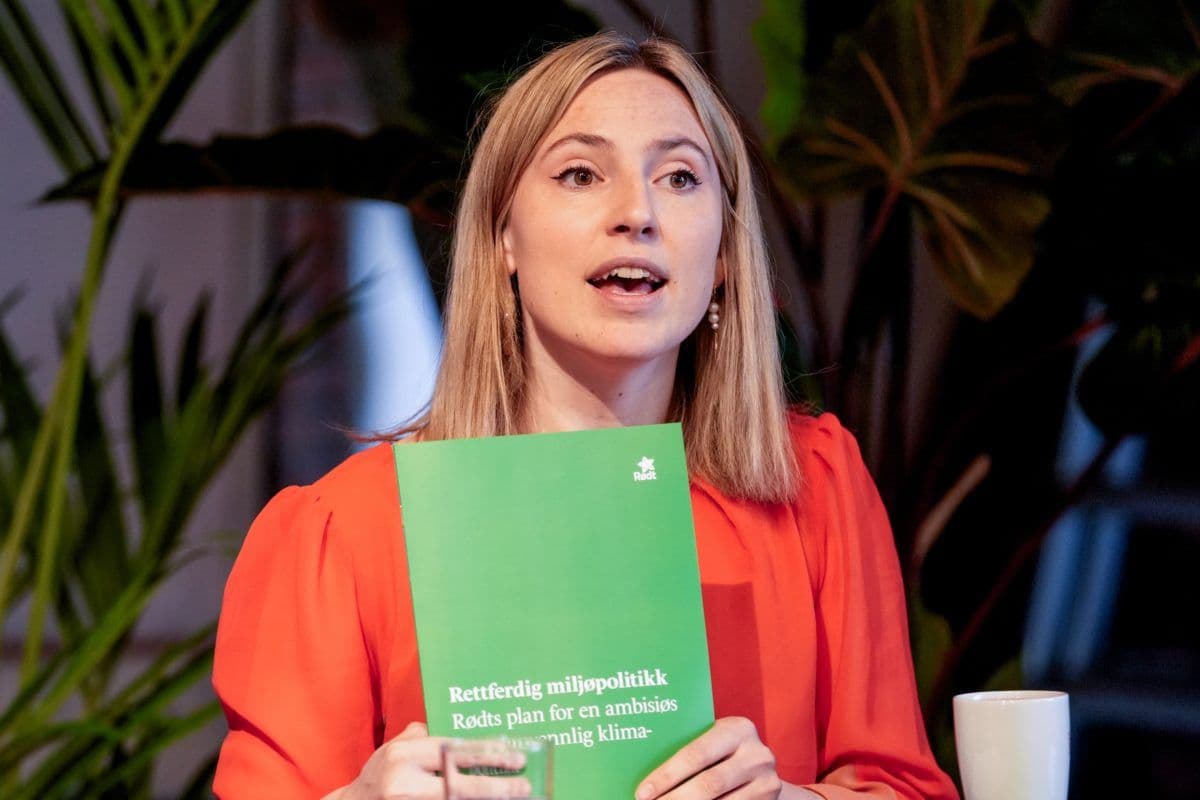 En kvinne i rød topp holder opp et grønt hefte med tittelen «Rettferdig miljøpolitikk» mens hun snakker innendørs, med planter i bakgrunnen.