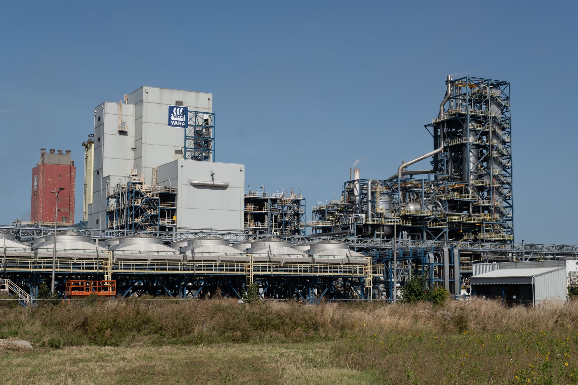 Et stort industrianlegg med en rekke sammenkoblede bygninger, tårn og lagertanker, sett fra et jorde under en klar, blå himmel.