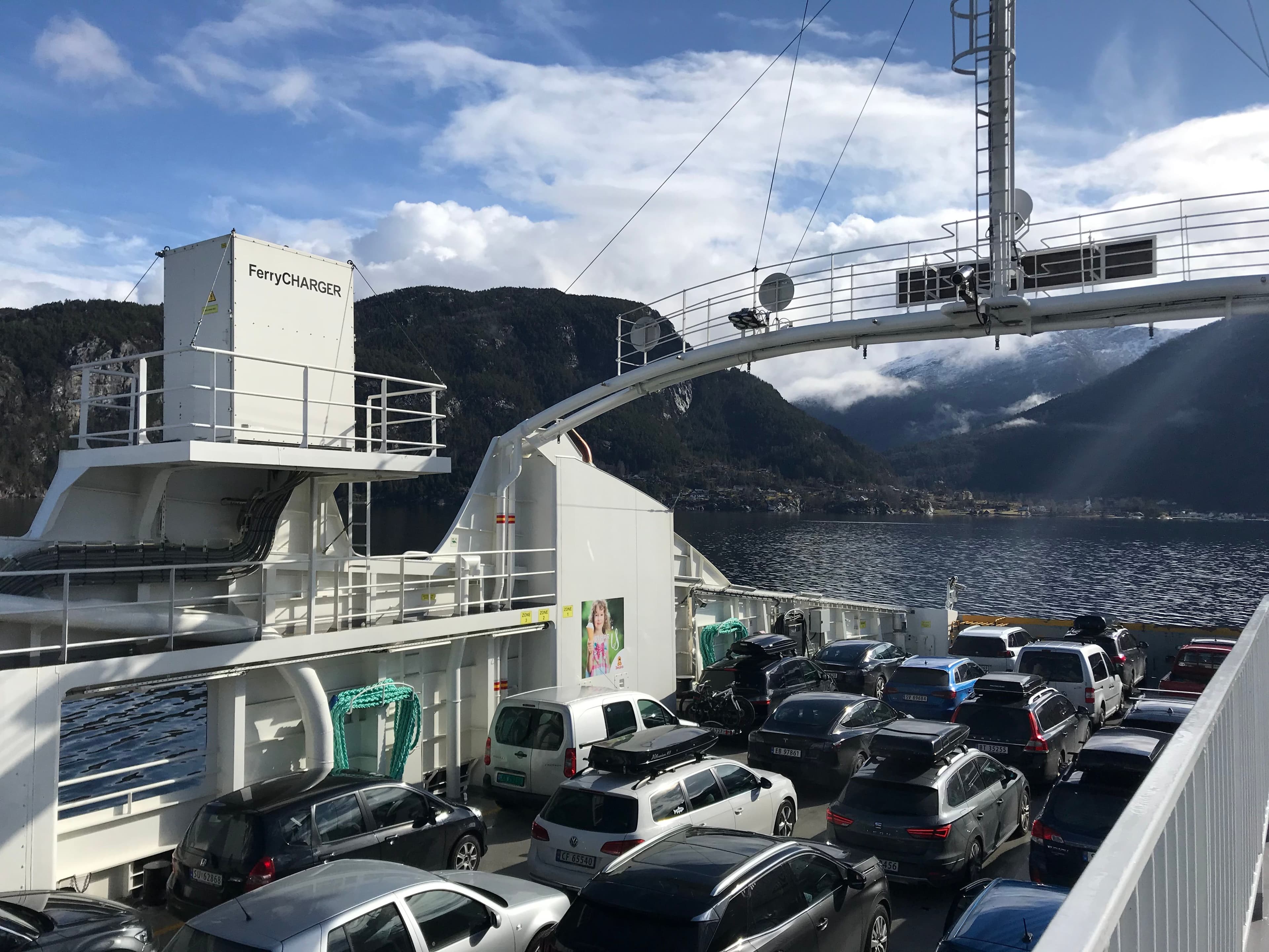 Ferjedekk fullt av biler og en «FerryCHARGER»-enhet, med fjell og vann i bakgrunnen.