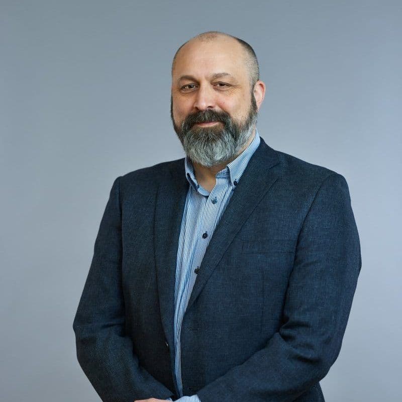 En mann med skjegg står foran en grå bakgrunn, iført mørk blazer og lyseblå stripete skjorte.