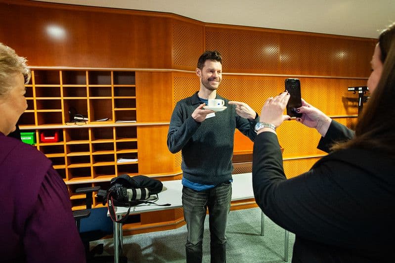 En smilende mann holder en kaffekopp og peker på den mens en annen person tar bilde av ham med en smarttelefon.