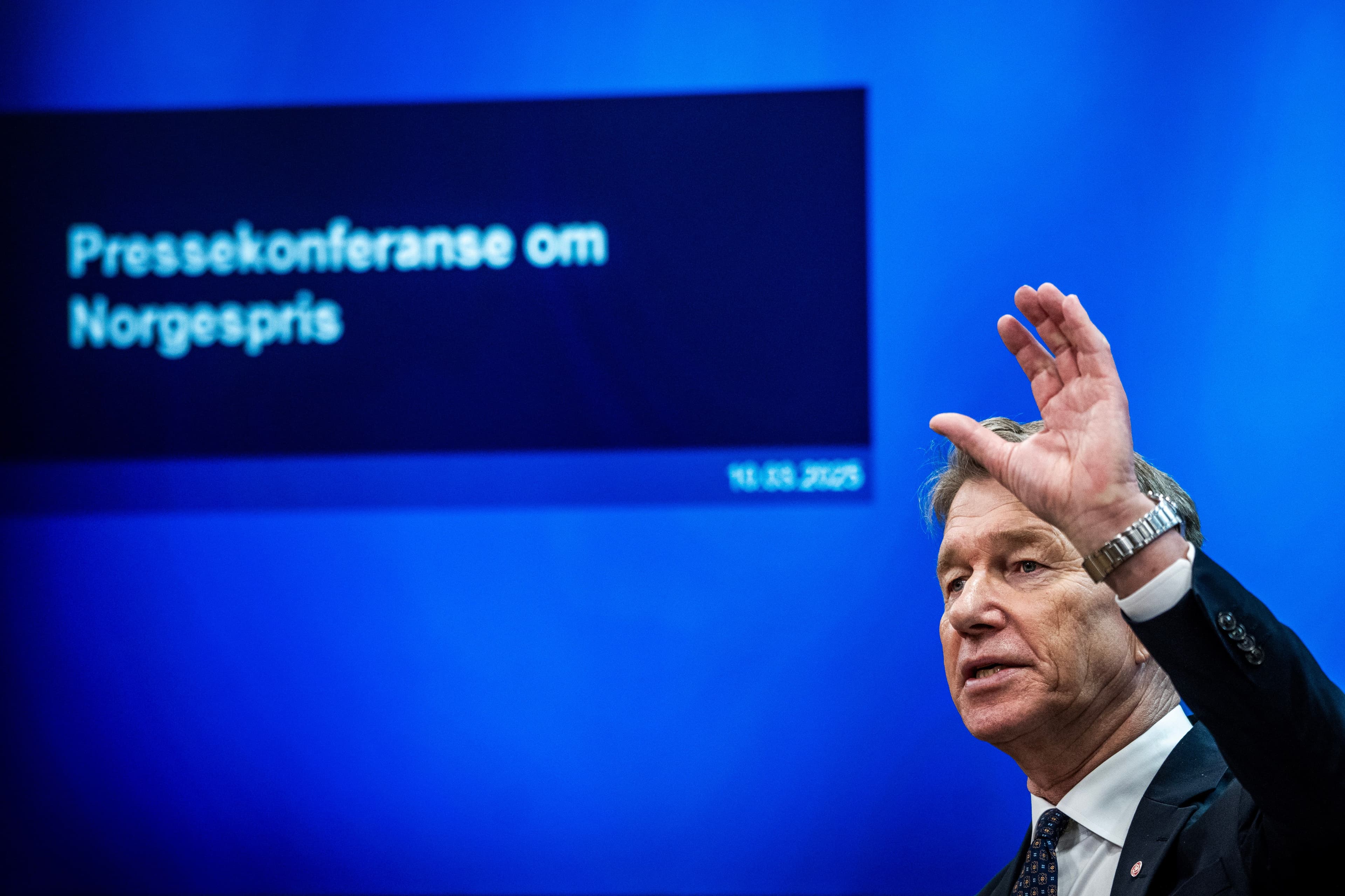 Mann ved en talerstol med hånden hevet, som snakker foran en blå skjerm med teksten «Pressekonferanse om Norgespris».