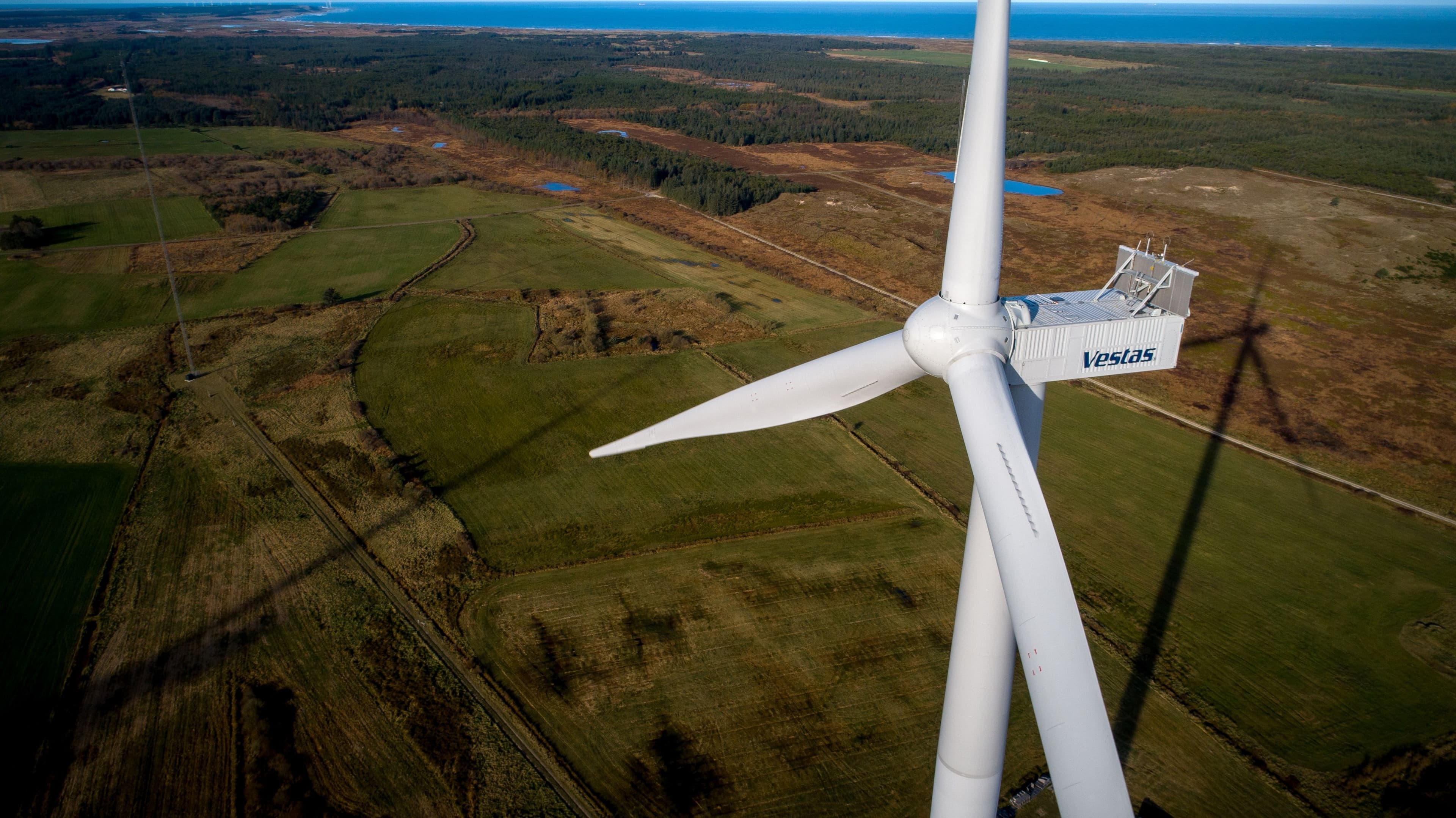 Fugleperspektiv av en Vestas-vindturbin som kaster en lang skygge over grønne jorder og skog, med havet i det fjerne.