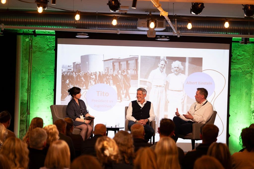 Tre personer sitter på en scene og diskuterer, med et projisert bilde av Tito og Albert Einstein bak seg. Et publikum sitter foran.