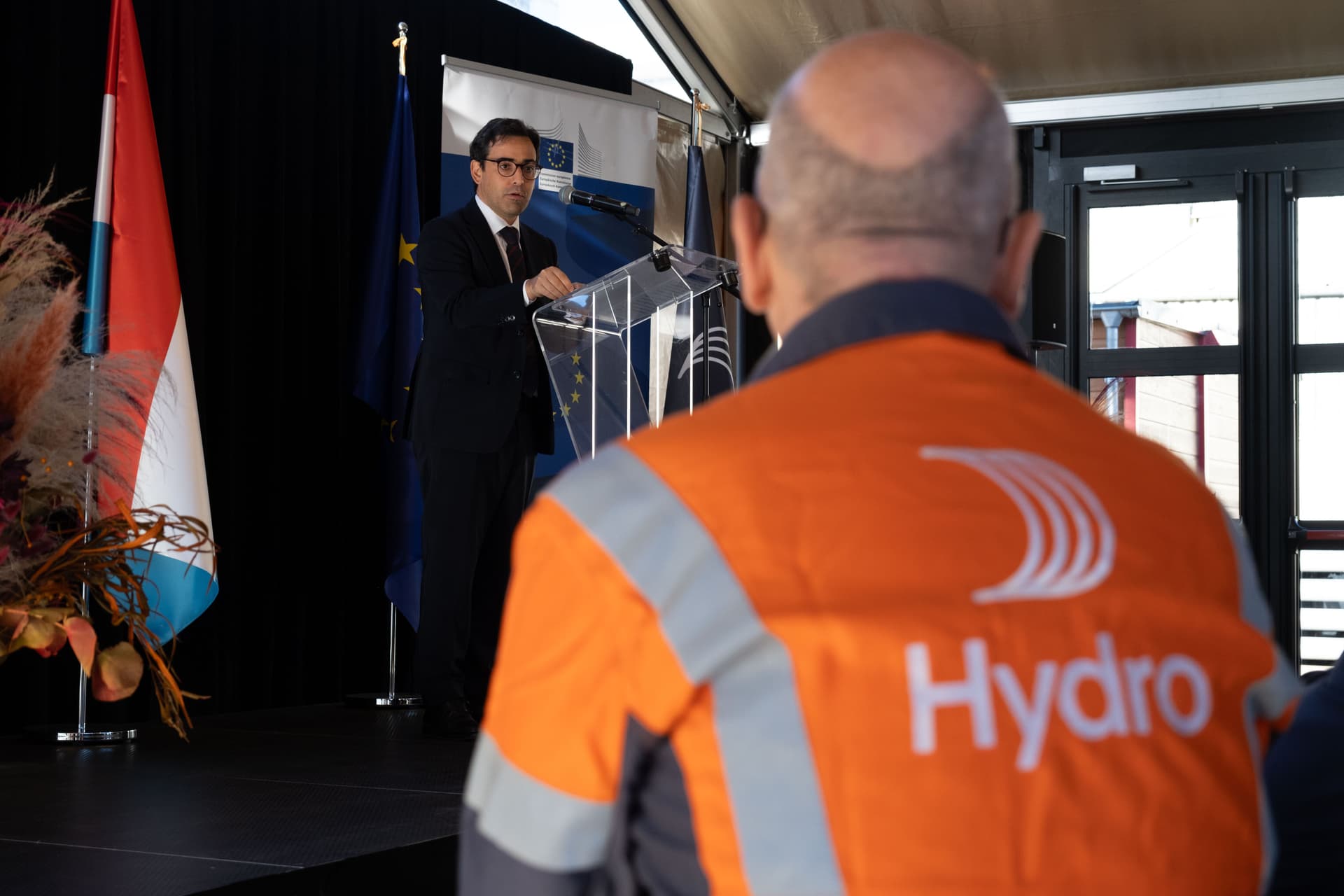 En mann i dress taler fra en talerstol, sett over skulderen til en person som har på seg en «Hydro»-sikkerhetsvest.