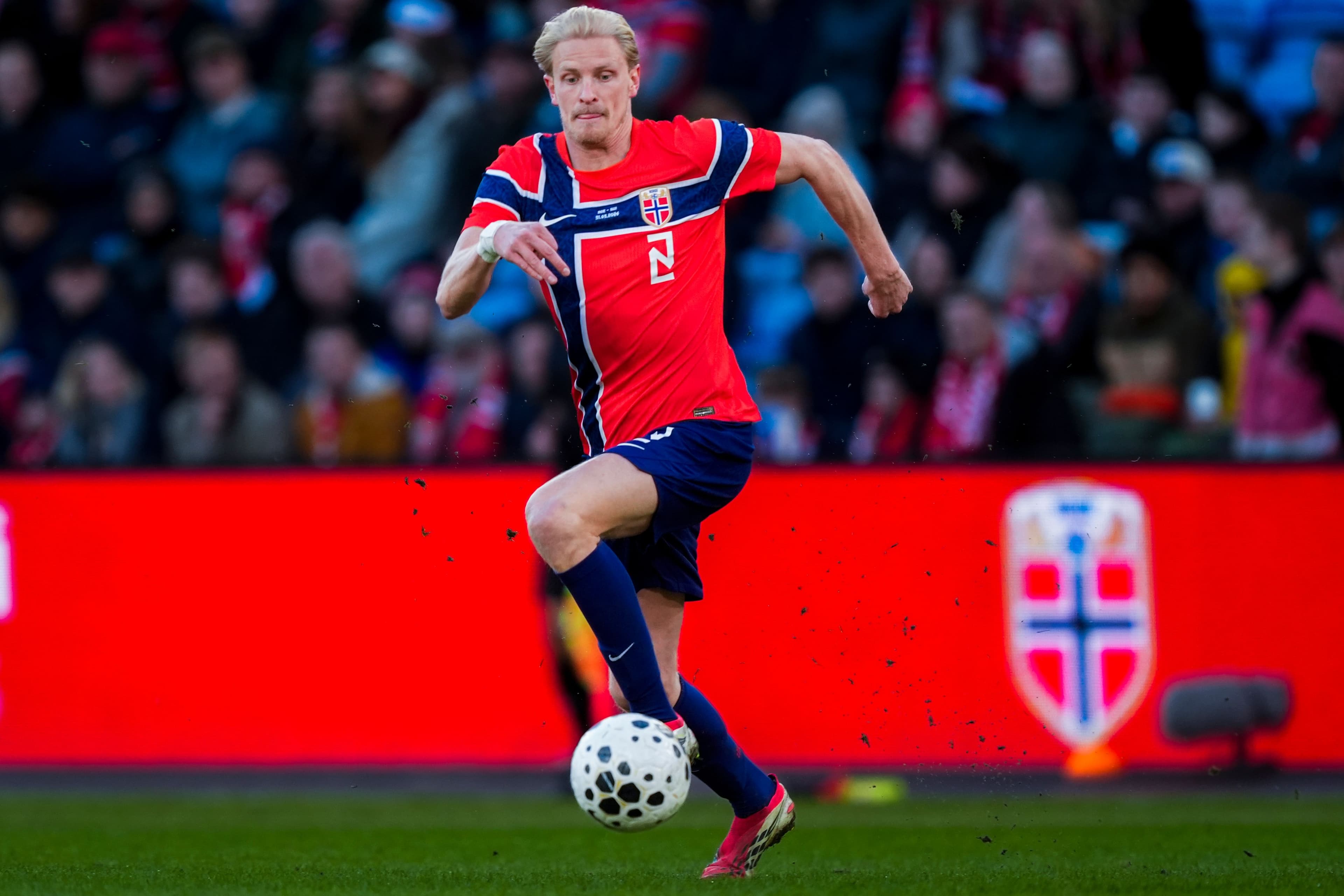 Blond norsk fotballspiller i rød og blå drakt som dribler en ball på en bane.
