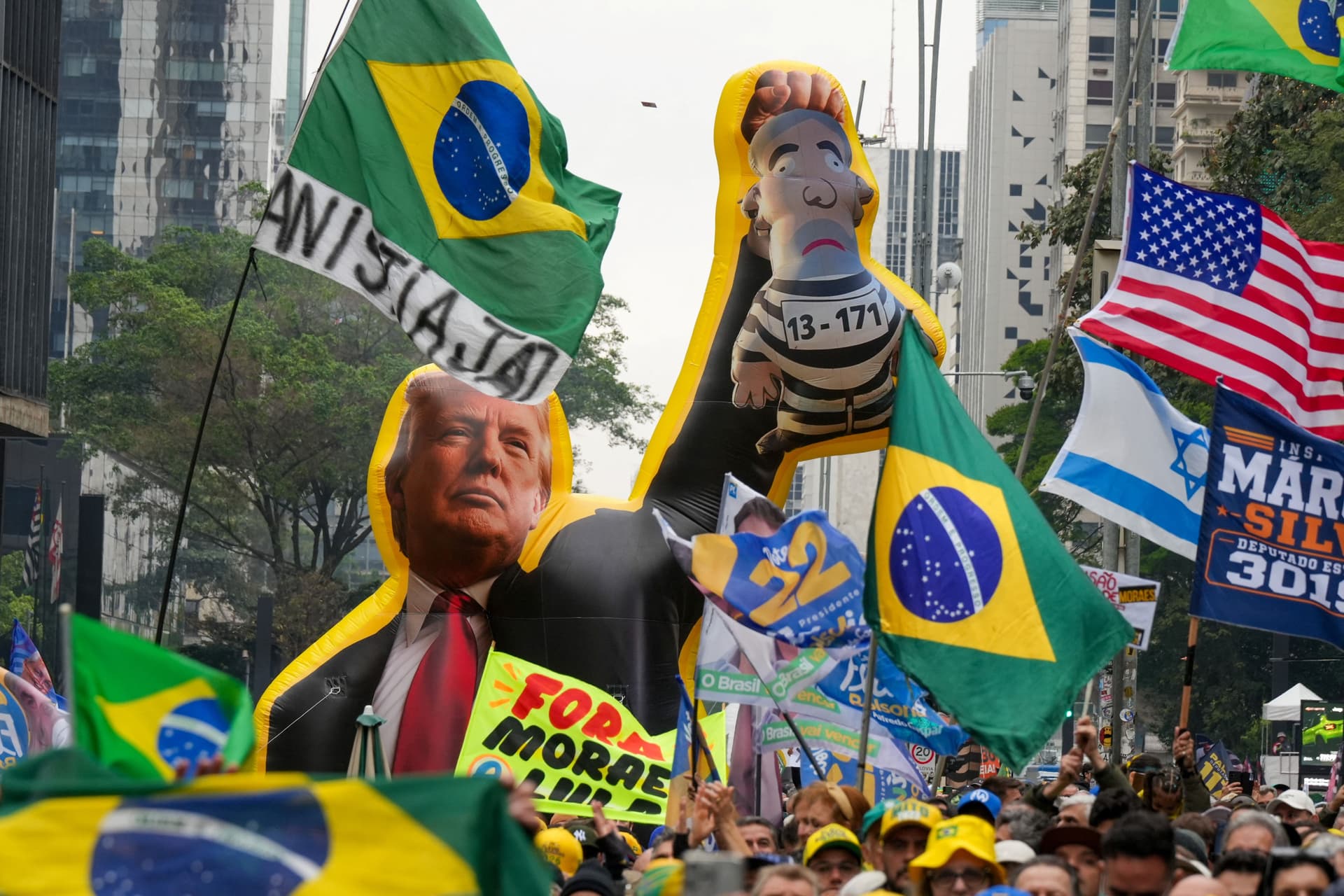 A street protest featuring large inflatables of Donald Trump and a figure in a prison uniform, surrounded by Brazilian, US, and Israeli flags, and signs including "FORA MORAES LULA."