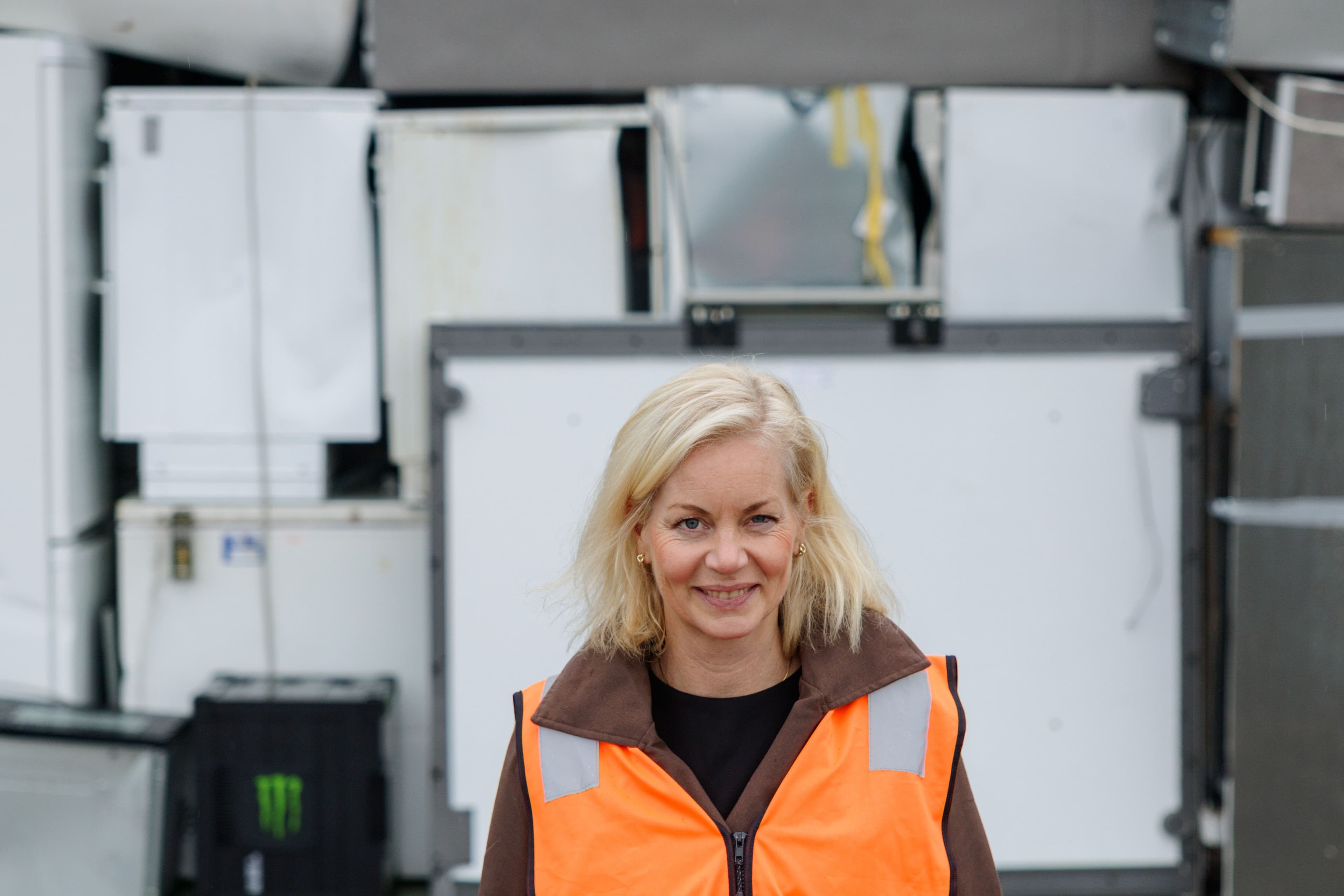 Smilende kvinne i oransje refleksvest foran stablede hvitevarer.