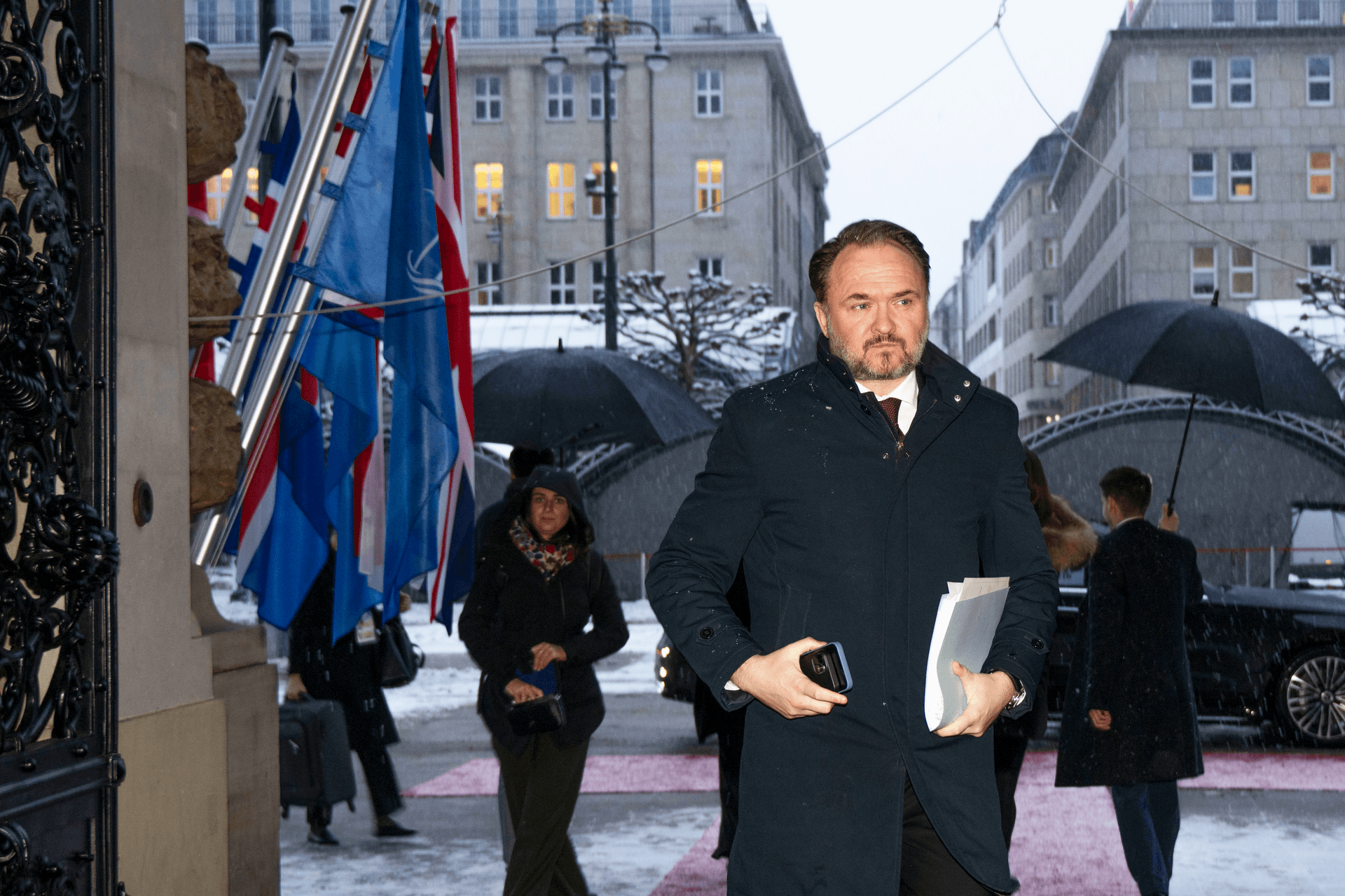 Mann i mørk frakk som går ut av en bygning med NATO- og andre flagg på en snørik dag.